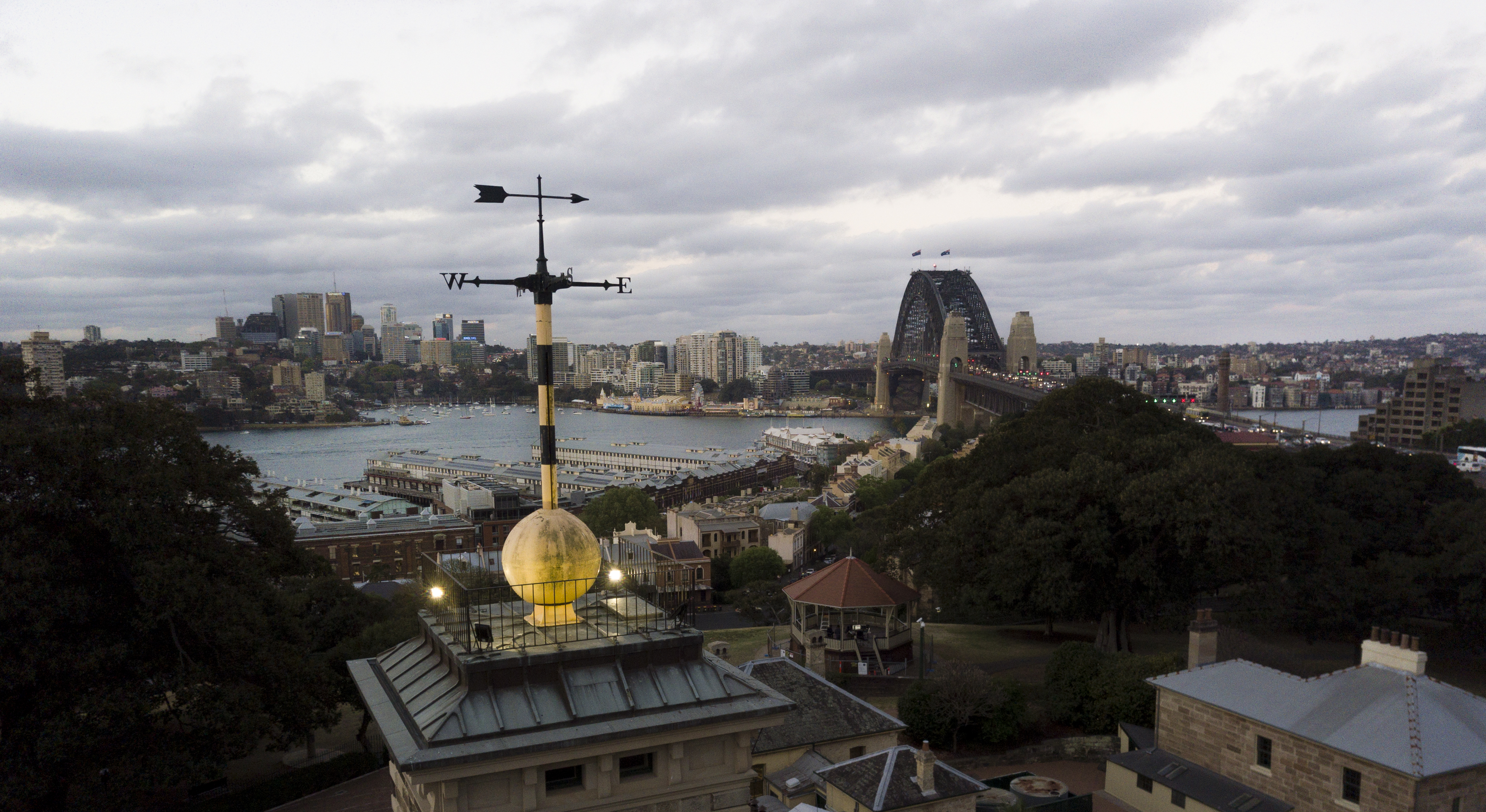 Aerial photo with a weathervane, mounted on top of a large yellow time ball, with various sandstone buildings below. The Sydney Harbour Bridge, The Rocks area, Sydney Harbour and Luna Park can be seen in the background.